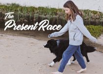 Cover of the album 'The Present Race' of a young girl and a dog walking on a beach.